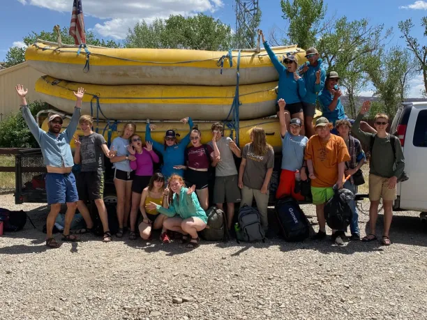 a group of students and mentors celebrate with a group photo in front of big rafts after an outdoor education program