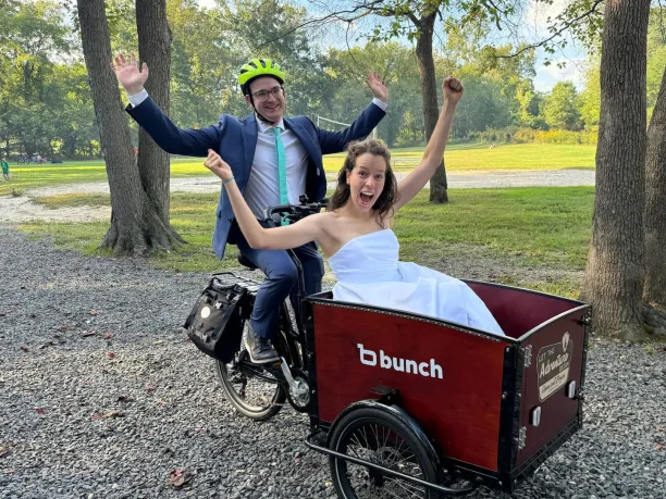 The groom is wearing a bright yellow helmet and sitting on the saddle of a cargo bike. The bridge is in the front basket of the bike. Both of them have their arms raised triumphantly