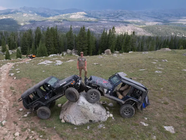 A man stands on top of two off-road vehicles parked on a large boulder. A vast mountain range extends behind him