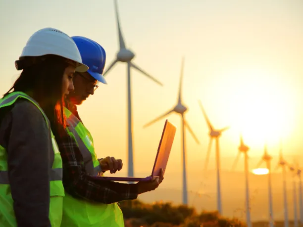 Two people wearing hard hats and reflective vests look at data on a laptop while the sun sets behind a row of wind turbines in the background