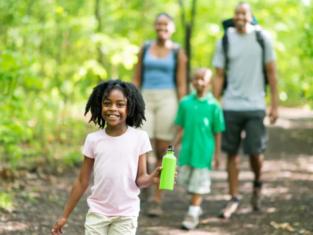 a little girl smiles while walking on a trail in the woods with her family close behind her