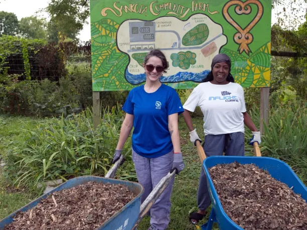 two women stand with wheelbarrels full of mulch in front of a sign for Sankofa Farm