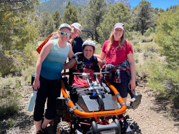 a woman sitting in an all-terrian wheel chair surrounded by her friends on a trail with mountains in the background
