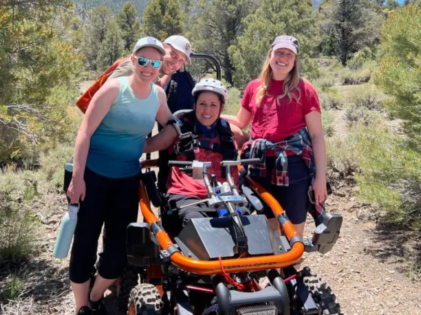 a woman sitting in an all-terrian wheel chair surrounded by her friends on a trail with mountains in the background