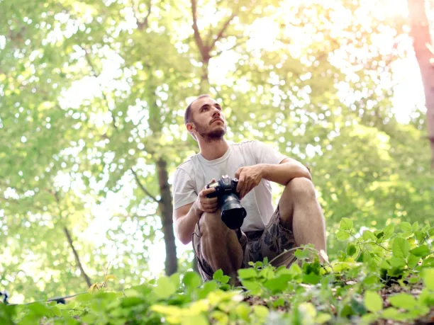 a man with a camera crouches in the forest while looking for birds