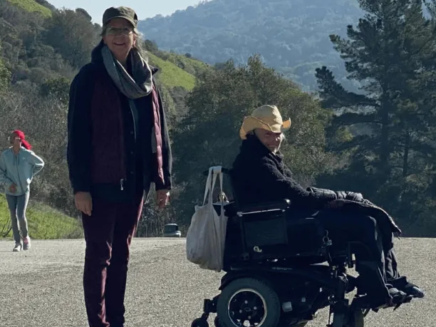 Two woman (one standing, one in a wheelchair) with big smiles hike Wildcat Creek Trail past green hills studded with trees 