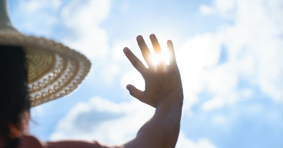 A person wearing a large sun hat holds their hand up to the sky to block the sun's harsh rays.