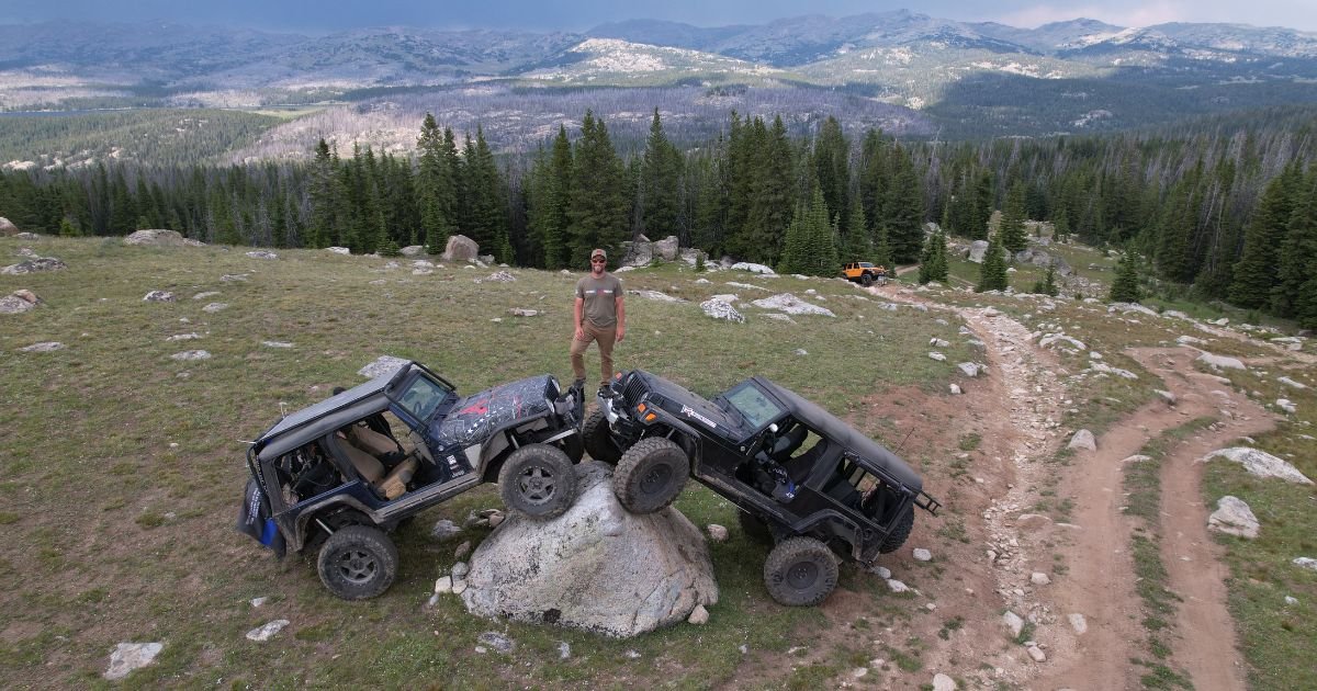 A man stands on top of two off-road vehicles parked on a large boulder. A vast mountain range extends behind him