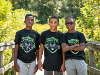 Three young black men stand together on a boardwalk in nature.