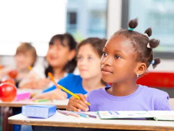 Young girl at desk holding pencil looking beyond frame at teacher