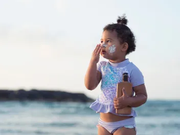 a little girl applies sunscreen to her face while standing on the beach