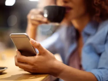 a woman holds a cell phone while reading and drinking from a mug