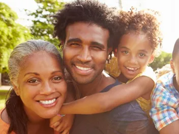 a black man holds his daughter on his back with his wife standing next to him all smiling