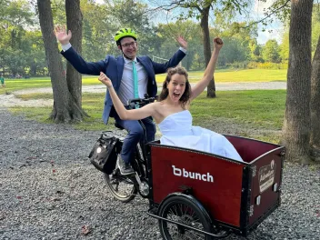 The groom is wearing a bright yellow helmet and sitting on the saddle of a cargo bike. The bridge is in the front basket of the bike. Both of them have their arms raised triumphantly