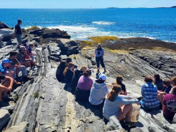 A woman stands on a rocky shoreline speaking to a group of seated college students as they overlook the ocean in the background