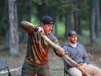 a man wearing camouflage stirs a bonfire while another man sits in a chair behind him in the forest