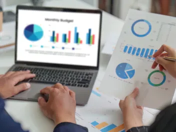 Two people's hands holding financial reports and typing on a laptop