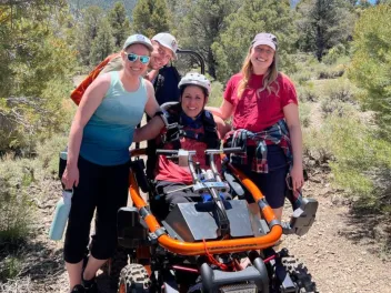 a woman sitting in an all-terrian wheel chair surrounded by her friends on a trail with mountains in the background