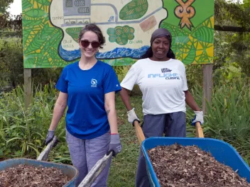 two women volunteers stand side by side holding wheelbarrows full of mulch