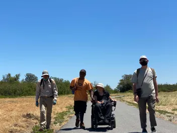 Three individuals, including one in a motorized wheelchair, hike with a park ranger on an asphalt path through dry grasslands at Big Break Regional Shoreline 
